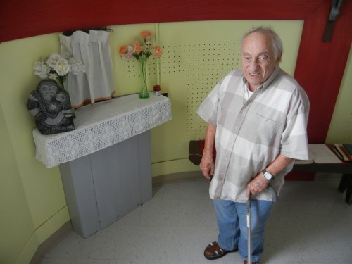 In June, 2013, Father Jules Dion stands in his mission near a carving made by Mitiarjuk Nappaaluk. (PHOTO BY JANE GEORGE)